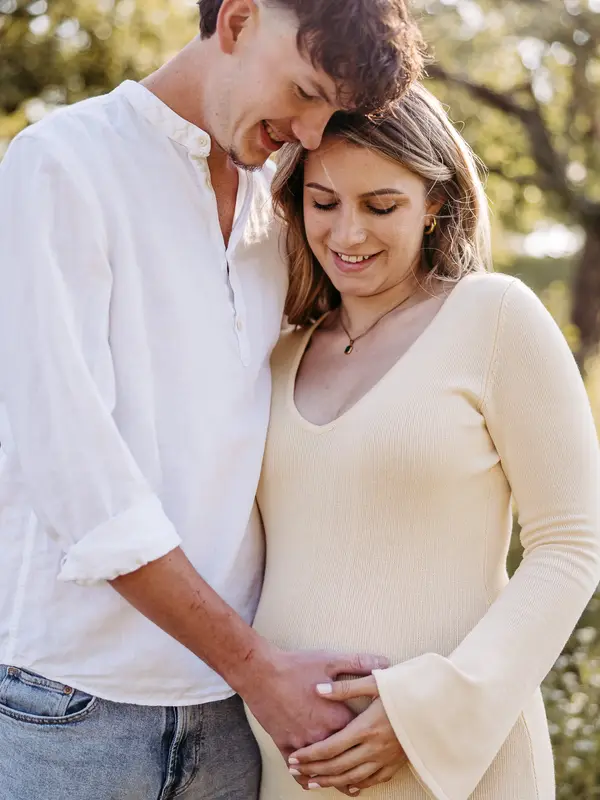 Paar im Abendlicht, er umarmt sie von hinten, ihre Hände auf dem Babybauch