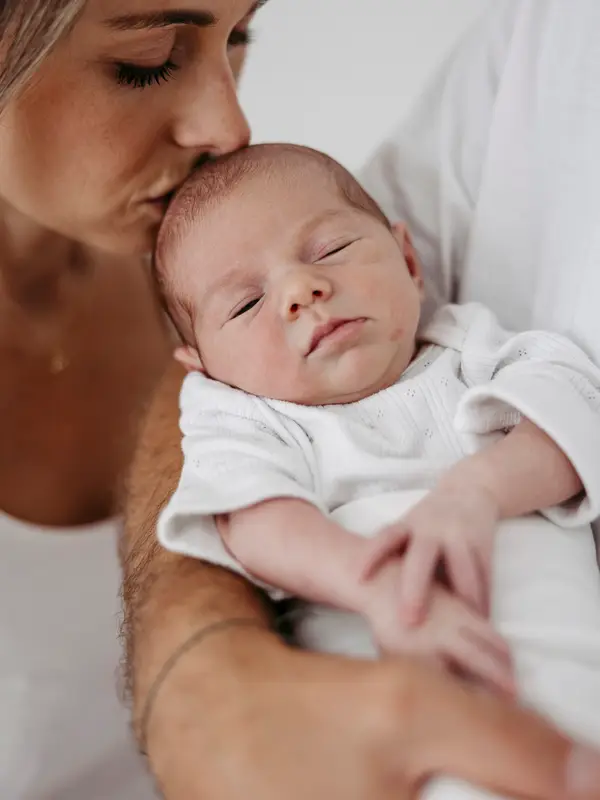Vater hält das schlafende Neugeborene, die Mutter küsst die Stirn des Babys