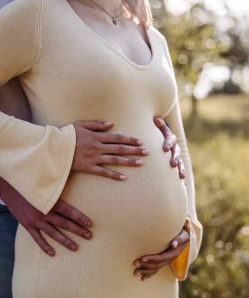 Hände eines Paares auf dem Babybauch, helles Strickkleid
